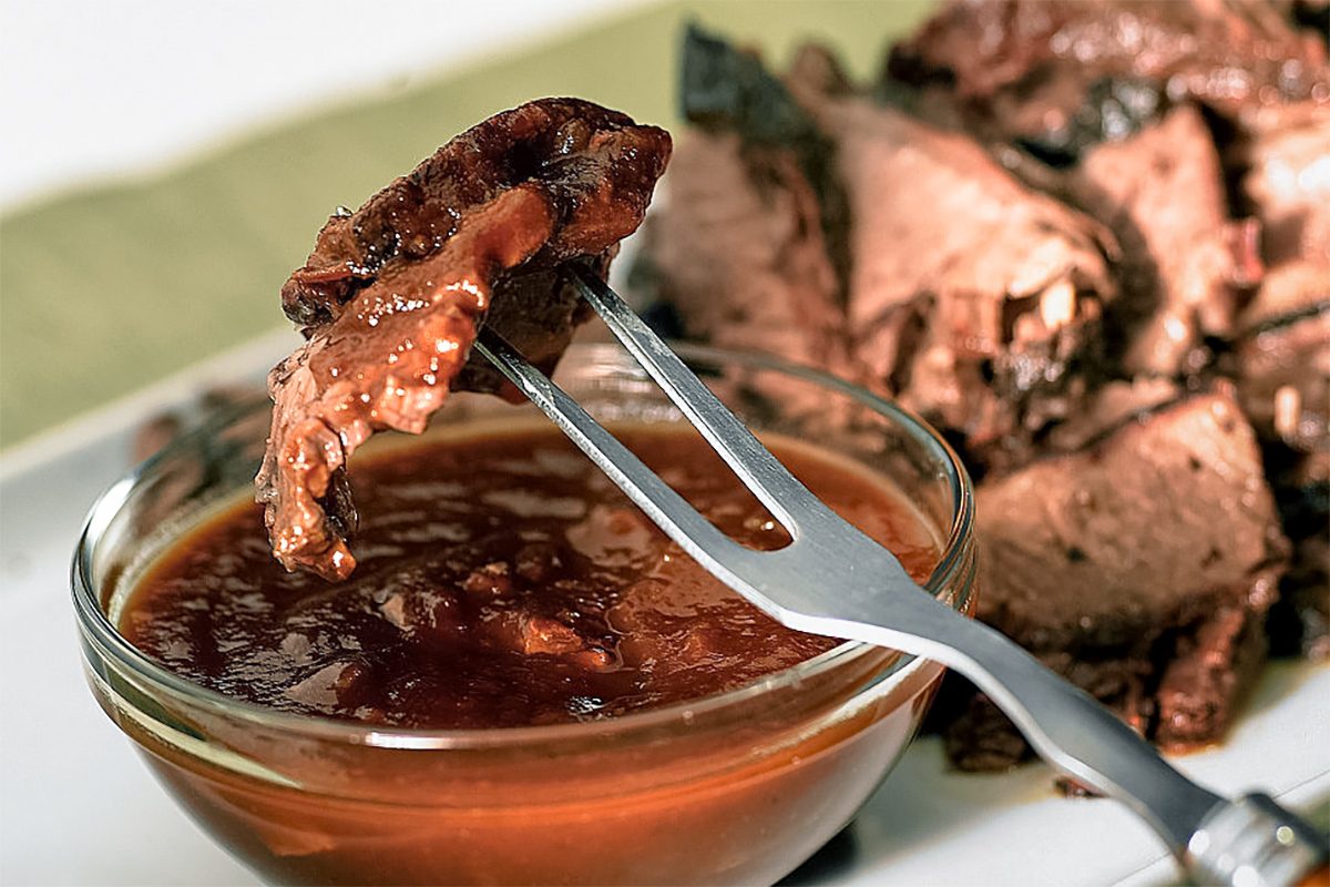 close up of a piece of smoked brisket on a fork over barbecue sauce in a glass bowl