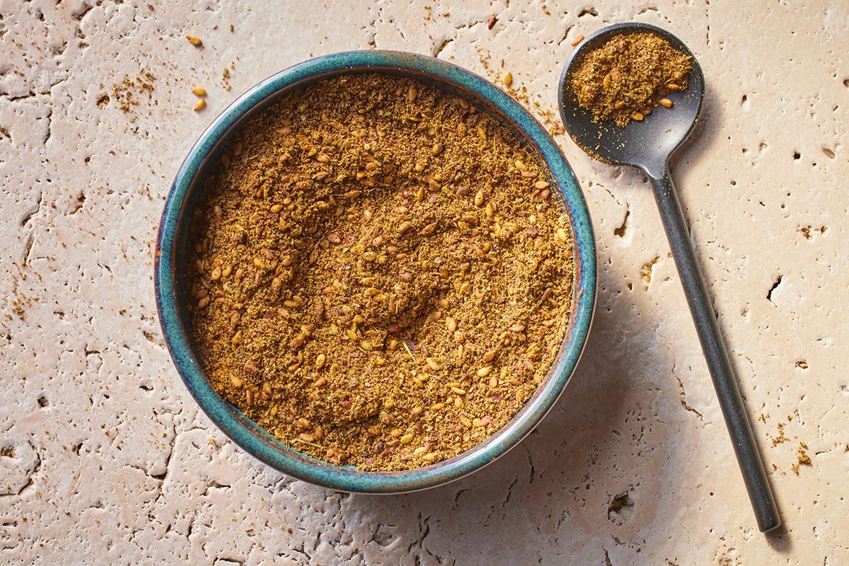 A blue bowl filled with a coarse, brownish spice blend sits on a light textured surface, with a metal spoon holding some of the spice mix beside it.