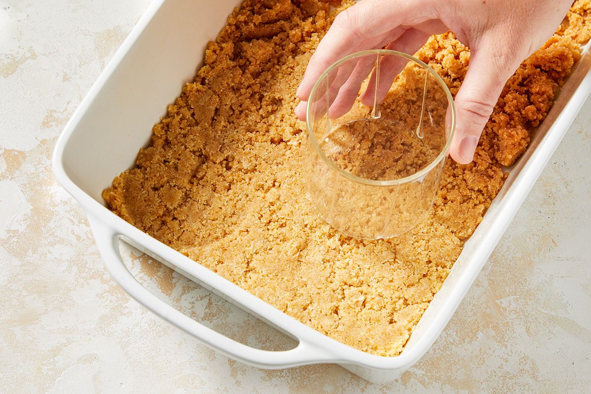Overhead horizontal image of pressing down graham cracker crumbs in a white baking dish with a measuring cup. The graham cracker mixture is pressed into the bottom of the dish to create a crust.