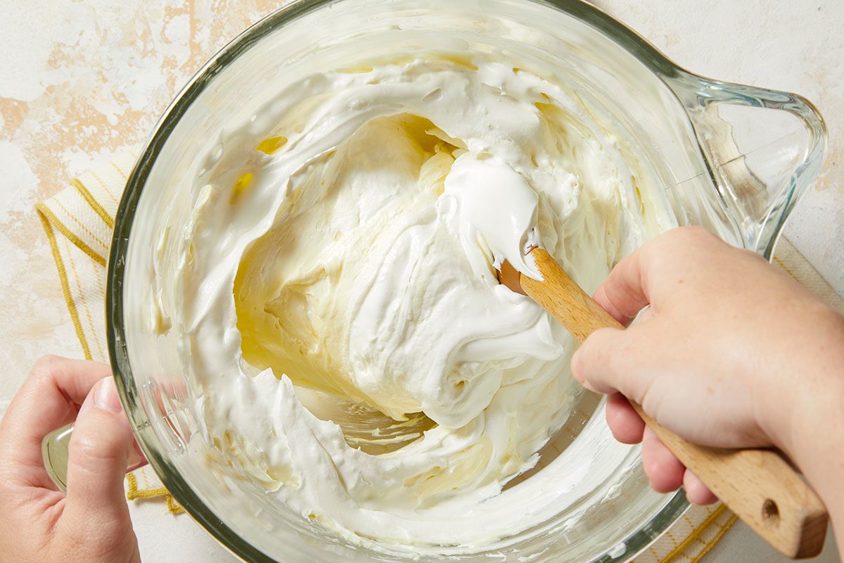 Overhead horizontal image of a clear glass mixing bowl with a thick, white mixture, possibly whipped cream or icing. A wooden spatula is being used to stir or fold the mixture. The bowl is being held by a hand.