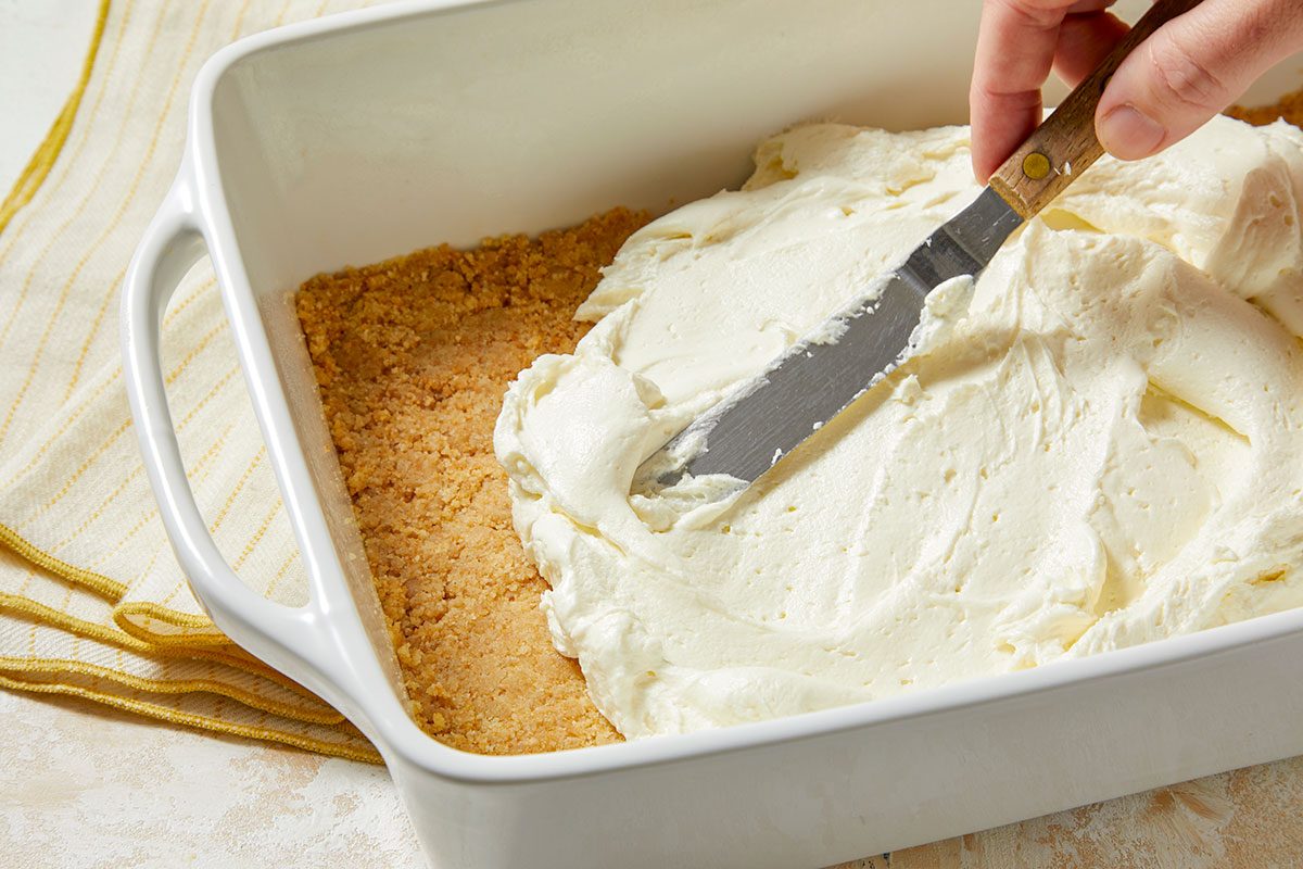 Eye Level horizontal image of a person spreading a thick layer of white cream filling onto a graham cracker crust in a white baking dish with a metal spatula. A striped yellow and white kitchen towel is partially visible to the side.