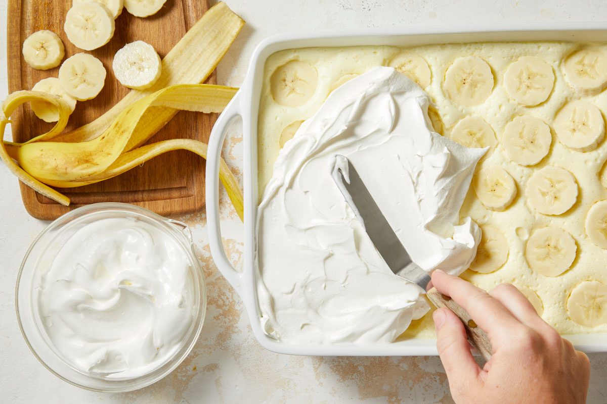 Overhead horizontal image of the application of frosting to a dessert with banana slices. A hand uses an offset spatula to spread a layer of fluffy white frosting on top of the dessert which is baking dish. Banana slices and a banana peel sit on a wooden cutting board.