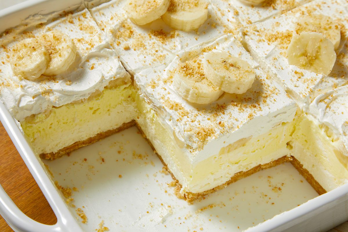 Eye Level horizontal image of banana pudding dessert cut into squares in a white baking dish. The slices are topped with whipped cream, sliced bananas, and crushed wafer crumbs.