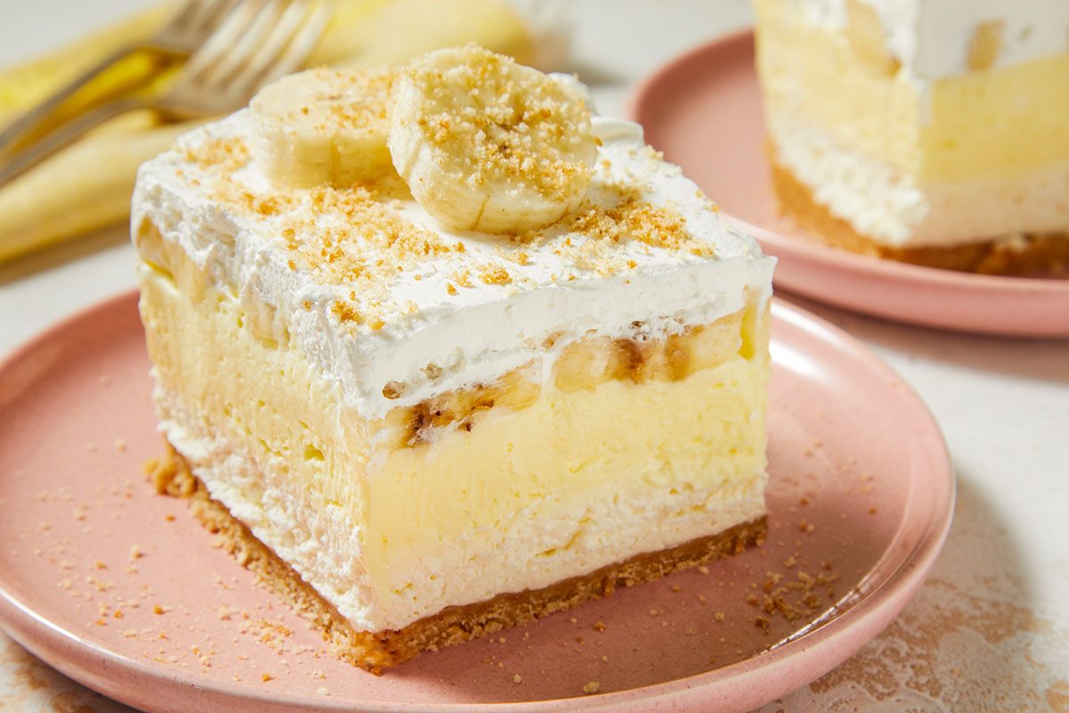 Eye Level horizontal image of a slice of banana pudding dessert on a pink plate. The slice is topped with whipped cream, banana slices, and graham cracker crumbs, and sits next to another slice of the same dessert, plus a fork.
