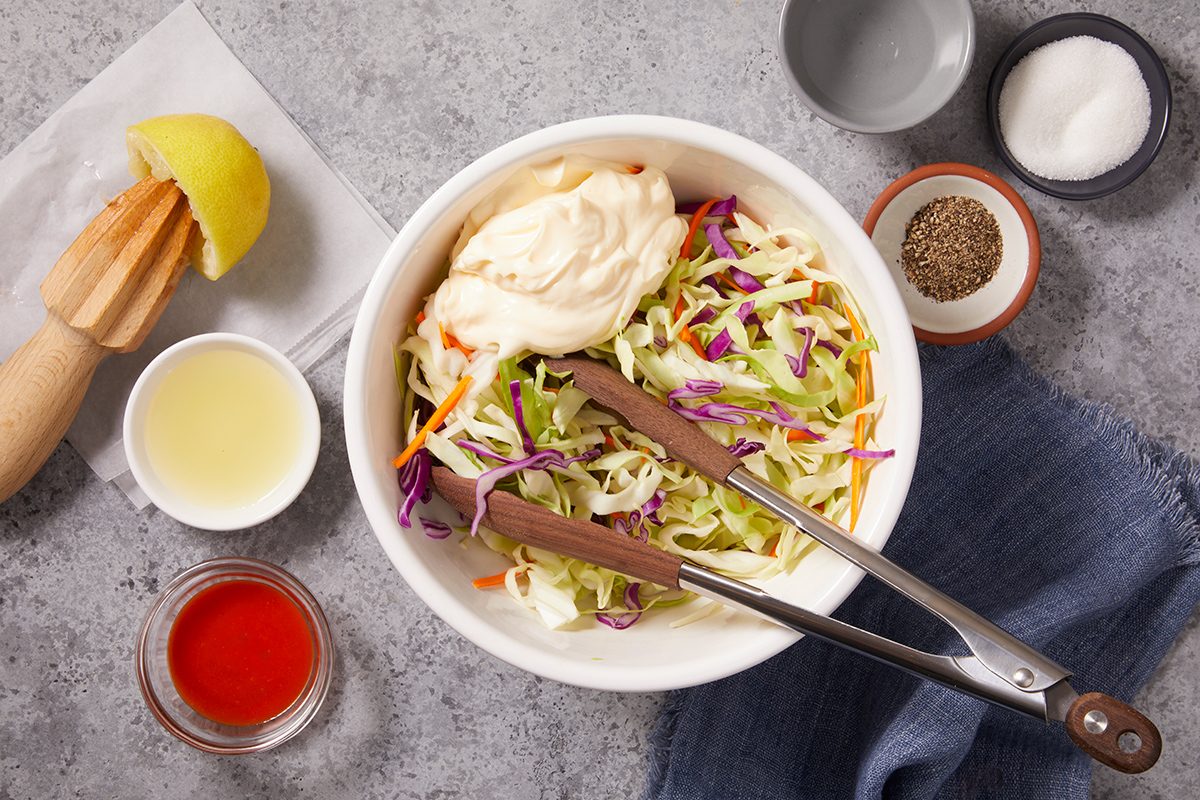 A bowl of coleslaw mix with mayonnaise and tongs, surrounded by small bowls of seasonings, lemon, juice, and dressing on a gray countertop.