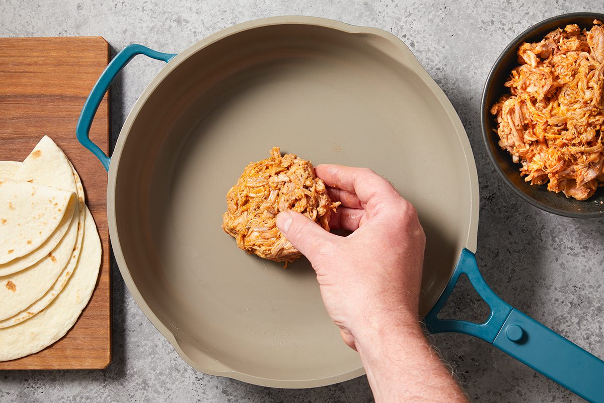 A hand places a portion of shredded chicken into a large skillet. Nearby are a stack of tortillas on a cutting board and a bowl filled with more shredded chicken.