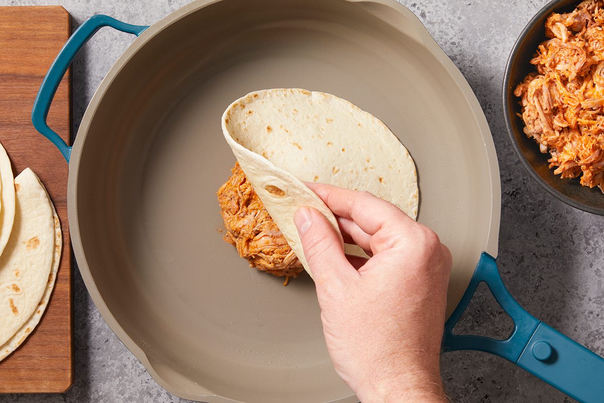 A hand folds a corn tortilla filled with shredded chicken inside a gray pan, with more tortillas and a bowl of chicken nearby.