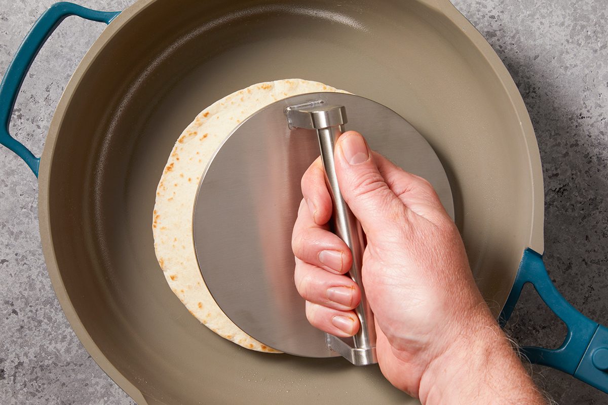 A hand presses a stainless steel tortilla press onto a tortilla inside a beige skillet with blue handles, on a gray countertop.