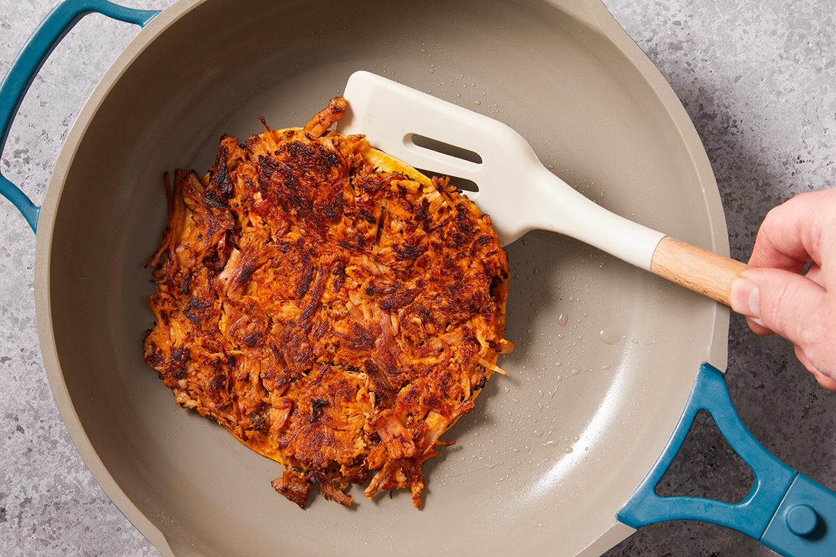A hand holds a white spatula, lifting a crispy, golden-brown hash brown from a light-colored skillet with blue handles on a gray surface.