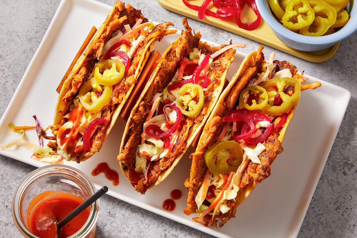 Three crispy tacos filled with shredded meat, cabbage, and topped with sliced pickled jalapeños and red onions, are arranged on a rectangular white plate. Sauces and extra toppings are in small dishes nearby.