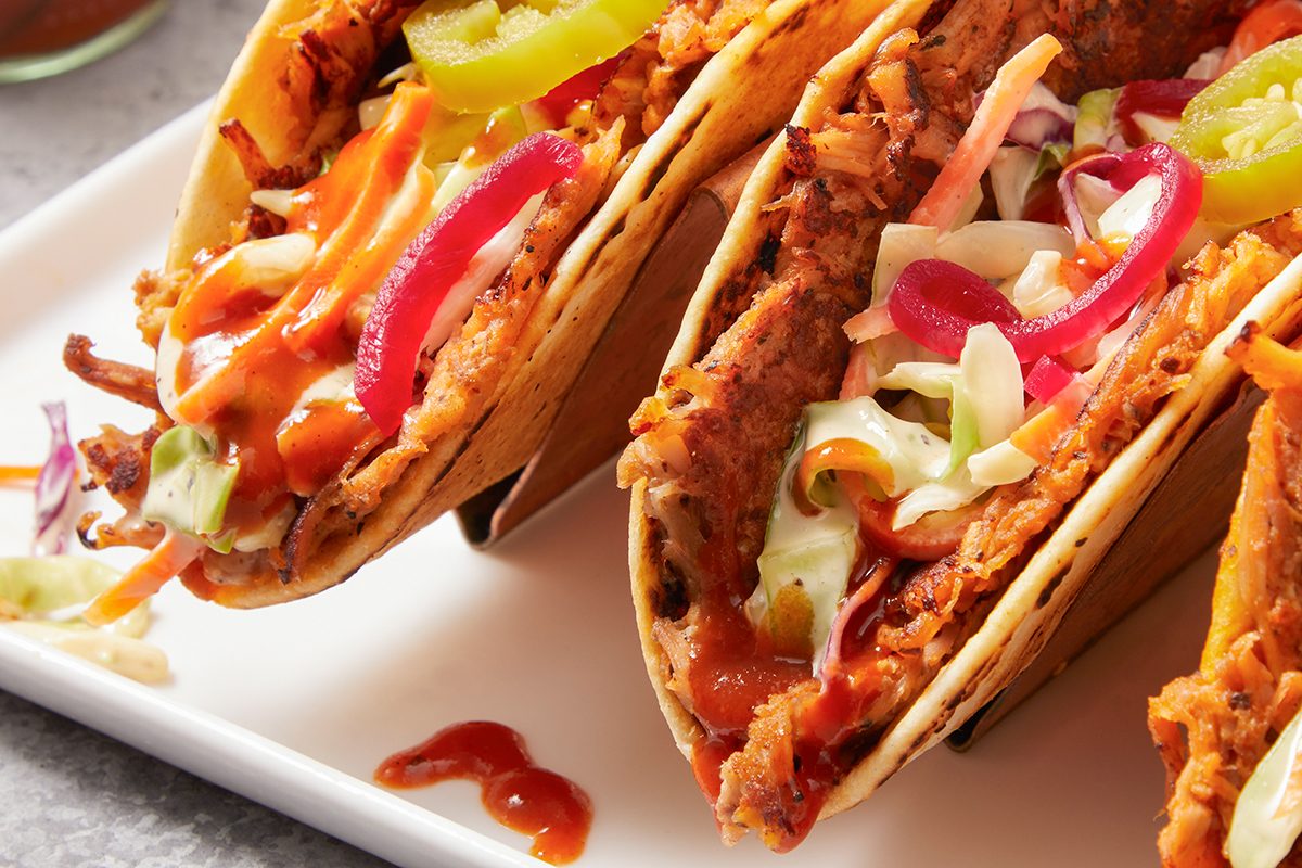 Close-up of two tacos filled with shredded meat, colorful slaw, sliced red onions, jalapeños, and drizzled with sauce, served on a white plate.
