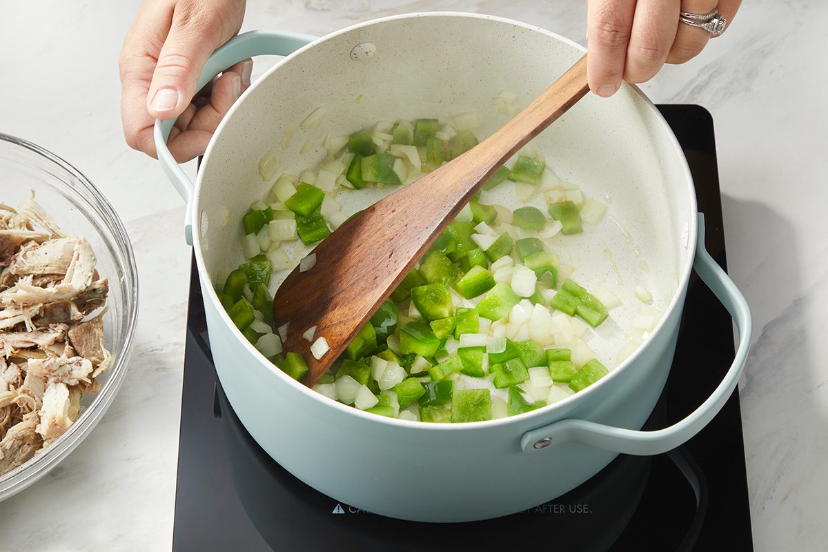 A person stirs chopped green bell peppers and onions in a light blue pot on a stovetop, with a bowl of shredded chicken nearby.