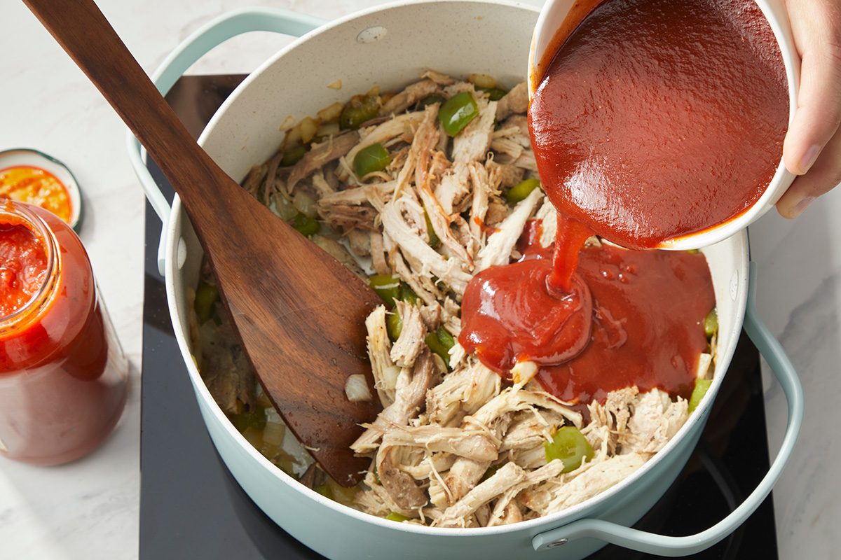 A hand pours tomato sauce from a bowl into a pot filled with shredded chicken, bell peppers, and onions, while a wooden spoon rests inside the pot.