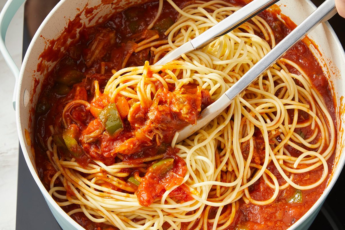 A pot of cooked spaghetti mixed with tomato sauce and vegetables, with metal tongs lifting a portion of pasta.