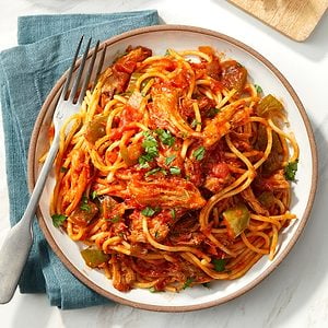 A plate of spaghetti topped with shredded meat, tomato sauce, chopped green bell peppers, and garnished with fresh herbs, served with a fork on a blue napkin.