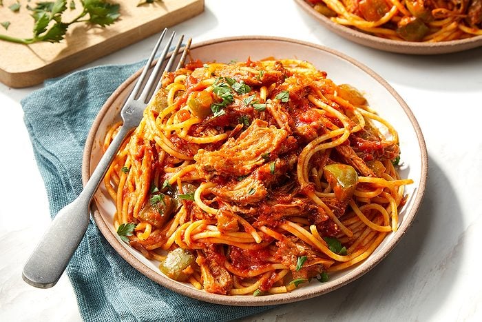 A plate of spaghetti with tomato sauce, shredded meat, and vegetables, garnished with herbs. A fork rests on the plate, and a blue napkin is underneath. Another plate and herbs are in the background.