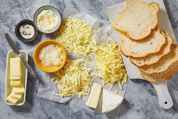 Overhead horizontal image of various ingredients prepared for a recipe, spread across a gray marble countertop. Slices of bread are arranged on a white cutting board, accompanied by piles of shredded cheese, a wedge of brie, bowls of mayonnaise, grated parmesan, and a white powder seasoning. Cubes of butter and a knife are also visible, laid out on parchment paper.