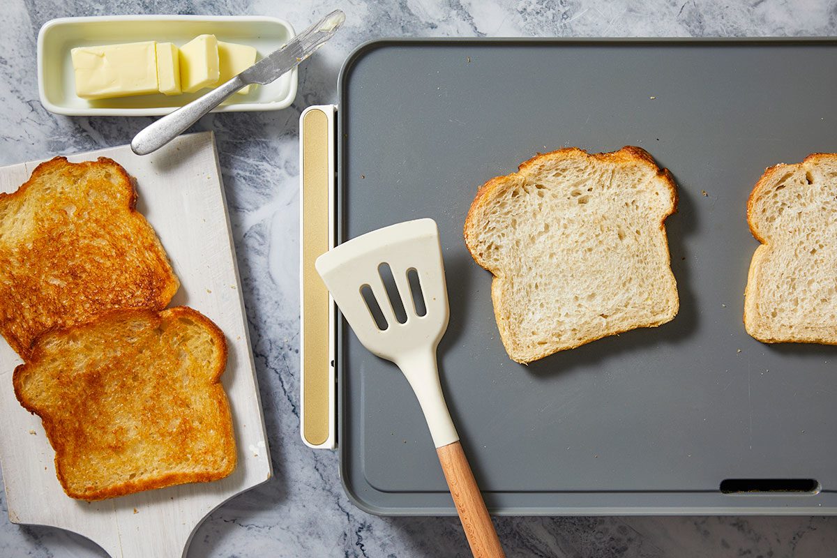 Overhead horizontal image of bread slices being prepared. Two slices of golden brown toast rest on a white cutting board, while two untoasted bread slices are on a gray electric griddle alongside a spatula. A dish of butter with a knife is also visible.