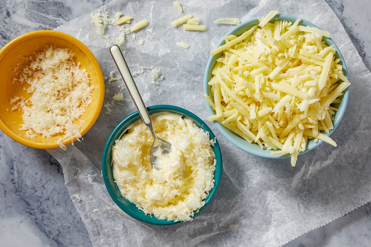 Overhead horizontal image of three bowls containing various cheeses and a creamy mixture, arranged on a piece of crumpled parchment paper. A yellow bowl holds finely grated cheese, a light blue bowl is filled with shredded yellow cheese, and a teal bowl contains a creamy white substance mixed with grated cheese and a spoon.