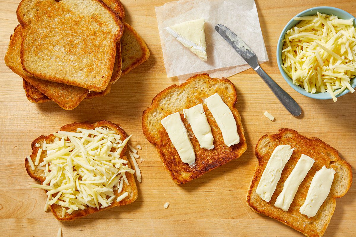 Overhead horizontal image of several slices of golden brown toast on a wooden cutting board. One slice of toast is topped with shredded cheese, while two others have long strips of soft white cheese on them. A bowl of shredded cheese, a triangular piece of brie, and a knife are also visible.