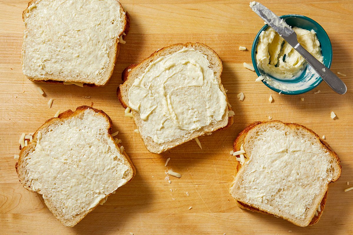 Overhead horizontal image of four slices of bread, each topped with a creamy spread. Shredded cheese is visible between the slices of bread, and a small blue bowl with additional spread and a knife rests nearby on the wooden cutting board.