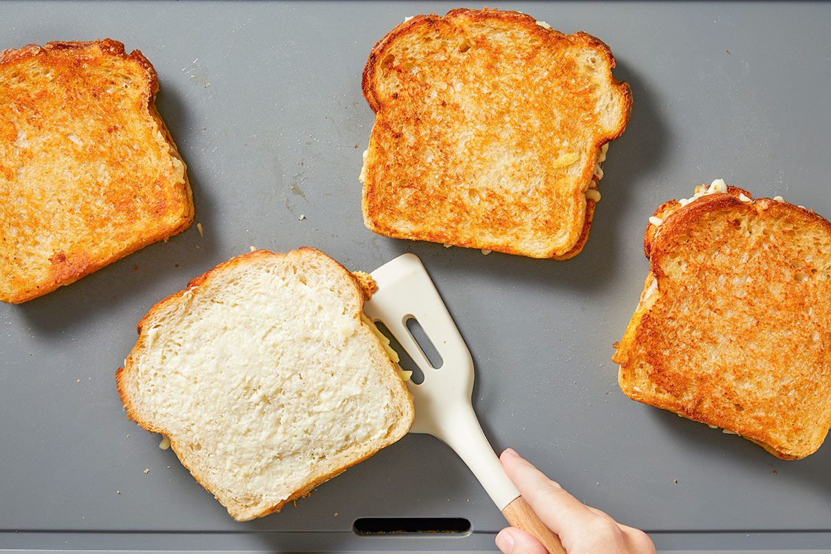 Overhead horizontal image of a hand using a white spatula to flip a grilled cheese sandwich on a gray electric griddle. Four sandwiches, made with bread, cheese, and a creamy spread, are cooking on the surface, with three already toasted golden brown.