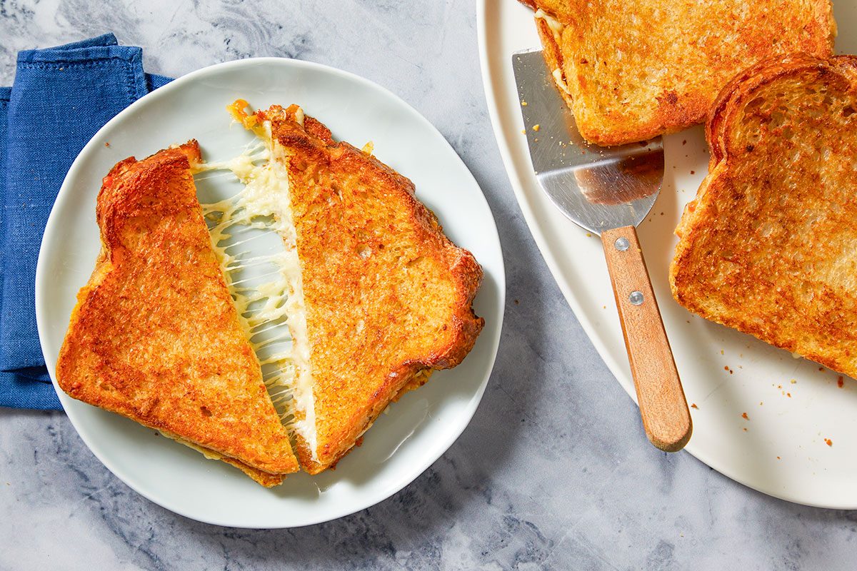 Overhead horizontal image of a golden-brown grilled cheese sandwich, cut in half on a white plate, showcasing a rich cheese pull with melted cheese stretching between the two halves. In the upper right corner, another whole grilled cheese sandwich is partially visible on a serving platter next to a metal spatula.