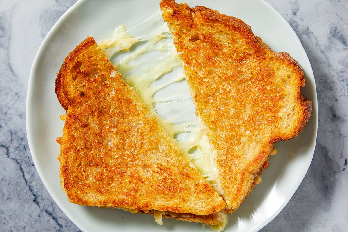 Overhead horizontal image of a grilled cheese sandwich cut in half, revealing gooey, stretchy melted cheese between two slices of golden-brown toasted bread. The sandwich rests on a light-colored plate against a grey marbled background.