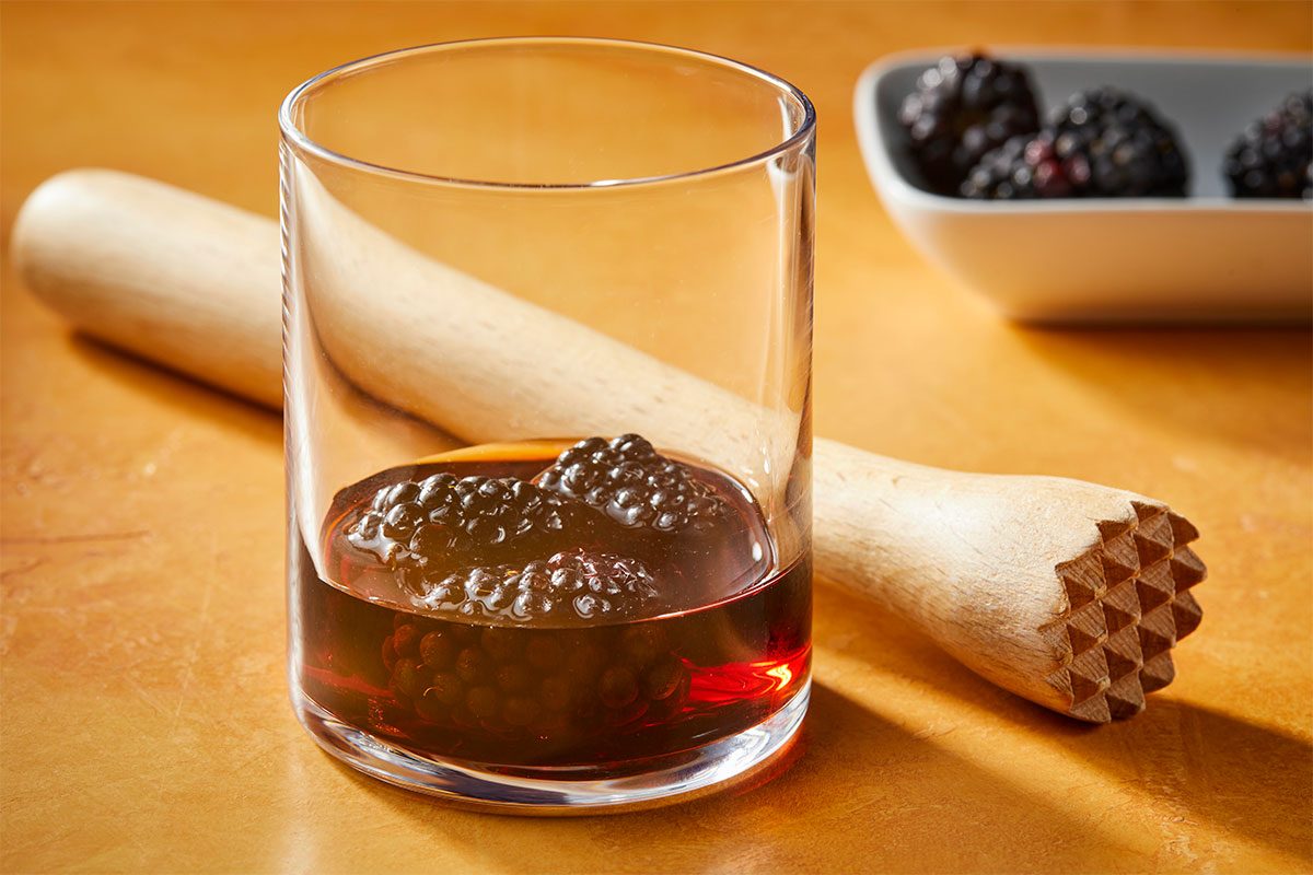 Eye Level horizontal image of blackberries and a dark liquid in a clear glass, ready to be muddled. A wooden muddler rests beside the glass on a light brown surface, with a white dish of additional blackberries blurred in the background. | [Original] RMS 283010 Blackberry Bramble; Trend Photography; How to; Step1; In a small glass, muddle blackberries and blackberry liqueur
