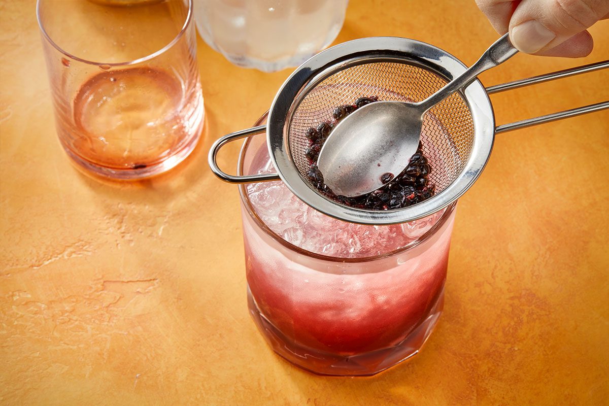 Eye Level horizontal image of a hand straining dark fruit pulp through a fine-mesh sieve into a glass filled with ice and a pink beverage. A spoon presses the fruit pulp to extract liquid into the drink. Another empty glass is visible to the left. | [Original] RMS 283010 Blackberry Bramble; Trend Photography; How to; Step 3; Fill a rocks glass three-quarters full with crushed ice. Strain the gin mixture into the glass. Add additional crushed ice; strain the blackberry liqueur mixture through a fine mesh strainer into the glass, lightly pressing with the back of a spoon as it strains. Garnish with a lemon wheel and a blackberry to serve