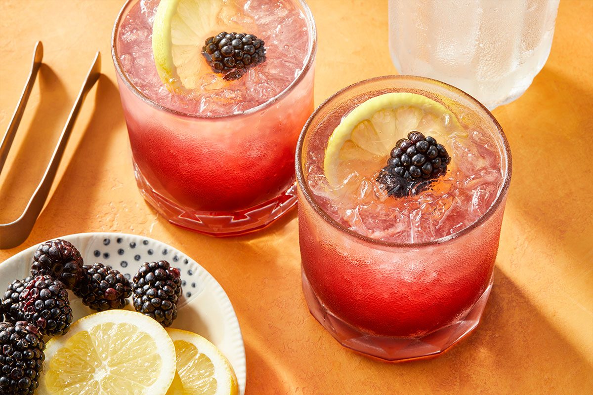 Eye Level horizontal image of two frosty glasses of red or purple cocktails with ice, garnished with a lemon slice and a blackberry. A small plate with additional blackberries and lemon slices, along with metal tongs, are visible on a textured orange surface, with another frosted glass in the background. | [Original] RMS 283010 Blackberry Bramble; Trend Photography; Horizontal AP; Pull