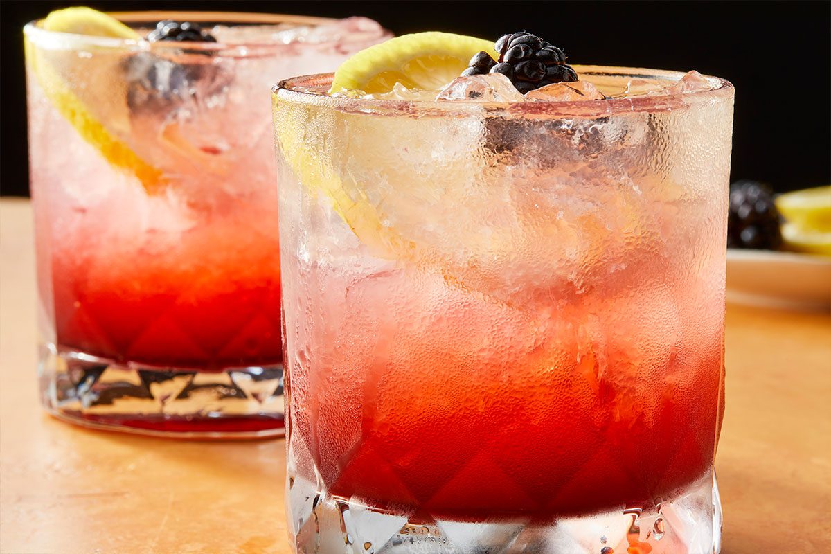 Eye level horizontal image of two frosted glasses filled with a vibrant red beverage. Each drink is garnished with ice, a lemon slice, and fresh blackberries. | [Original] RMS 283010 Blackberry Bramble; Trend Photography; Horizontal; Push