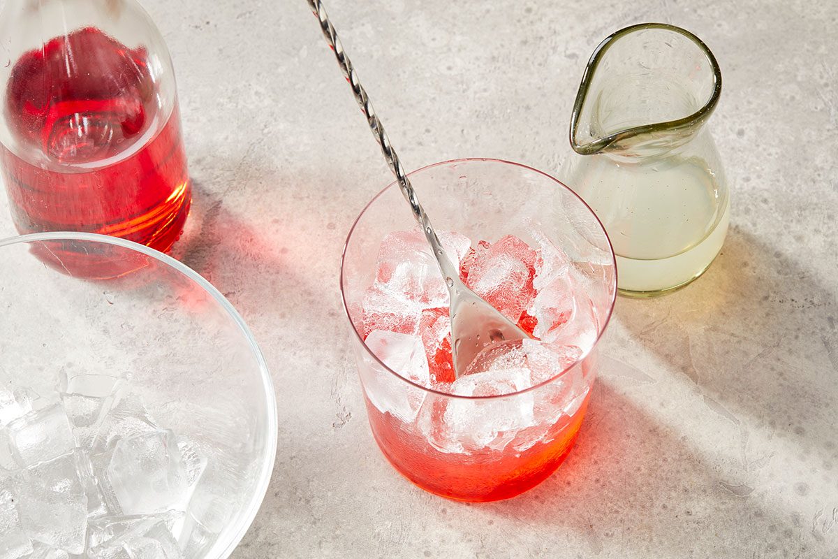 Overhead horizontal image of a clear glass containing red liquid and ice being stirred with a long metal spoon. A bottle of red liquid, a pitcher of light liquid, and another glass with ice are also visible.