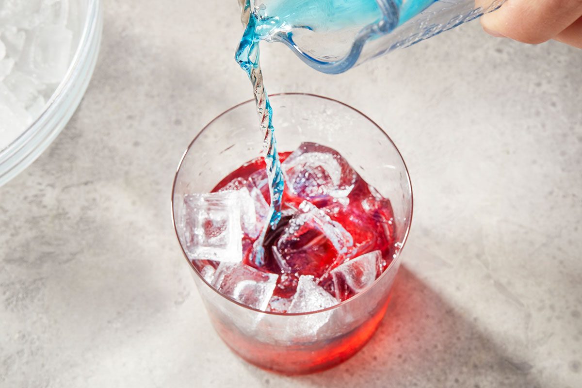 Overhead horizontal image of a hand pouring blue liquid from a clear measuring cup into a glass filled with ice and red liquid. More ice cubes are visible in a bowl in the background.