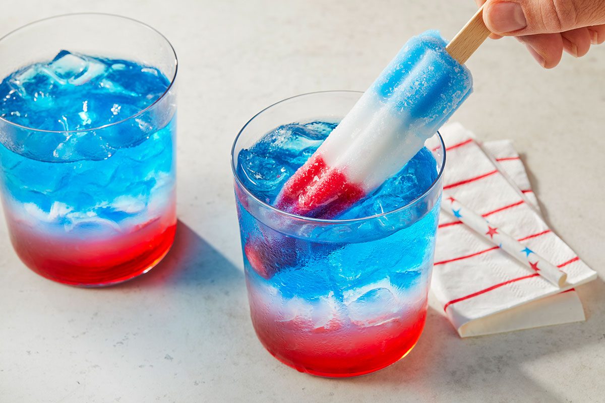 Eye Level horizontal image of a hand placing a red, white, and blue Bomb Pop into a glass filled with a layered red and blue drink and ice. Another similar drink is visible in the background, next to a patriotic striped napkin.