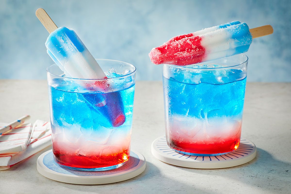 Eye level horizontal image of two glasses filled with a festive red, white, and blue layered drink, each featuring a Bomb Pop popsicle. One popsicle is partially submerged in the blue liquid, while the other rests on the rim of its glass, creating a vibrant, patriotic beverage.