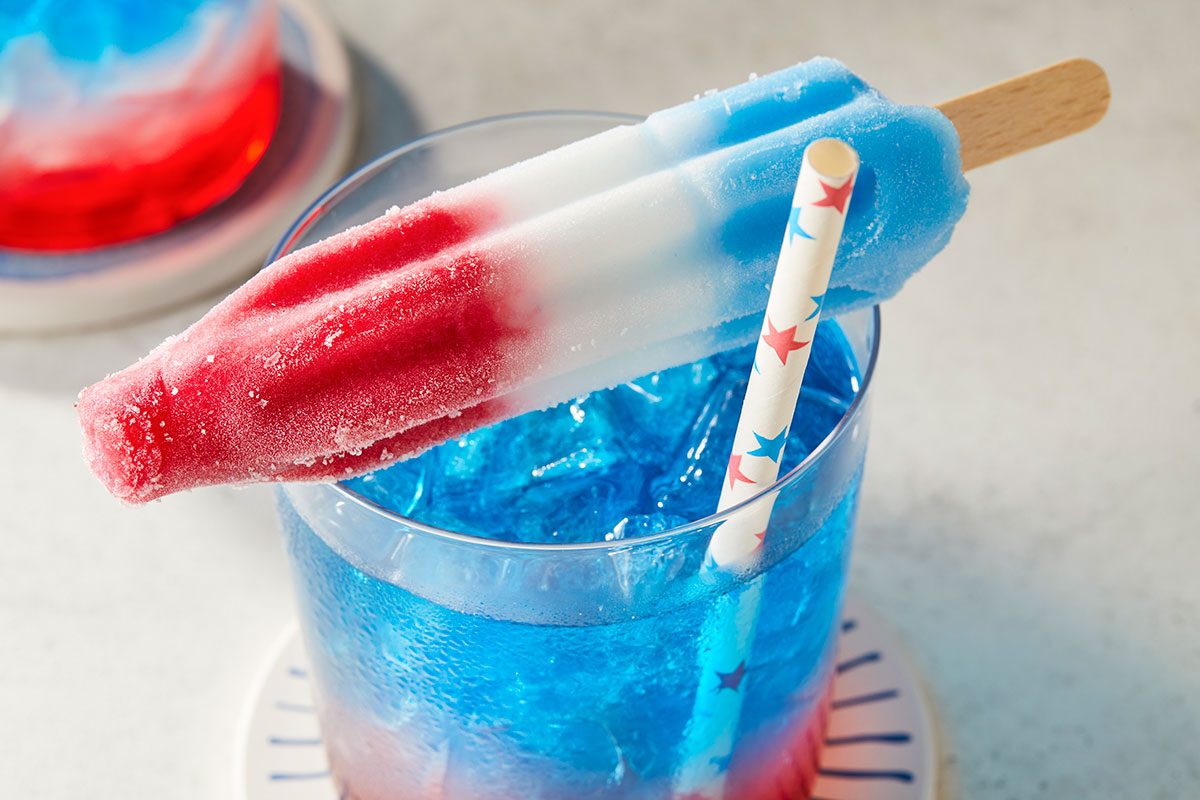 Eye Level horizontal image of a red, white, and blue rocket-shaped popsicle resting on the rim of a clear glass. The glass is filled with a vibrant blue beverage over ice, with a festive paper straw submerged in the drink. In the blurred background, another glass with a red and blue drink is visible, emphasizing a patriotic theme.