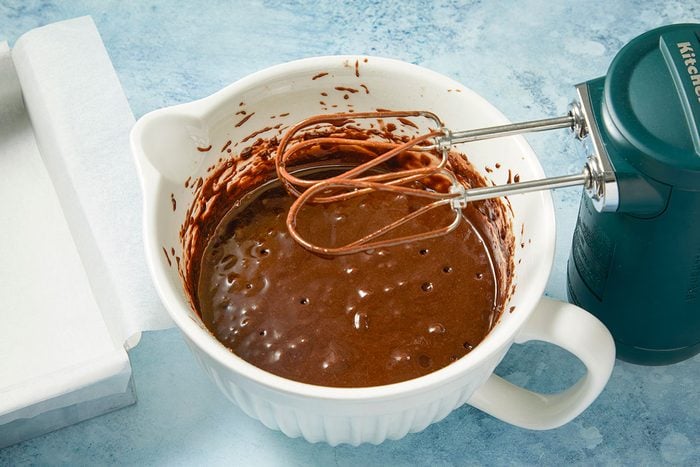 Overhead horizontal image of chocolate batter being mixed in a white bowl with a KitchenAid hand mixer. Also visible is parchment paper on the side.