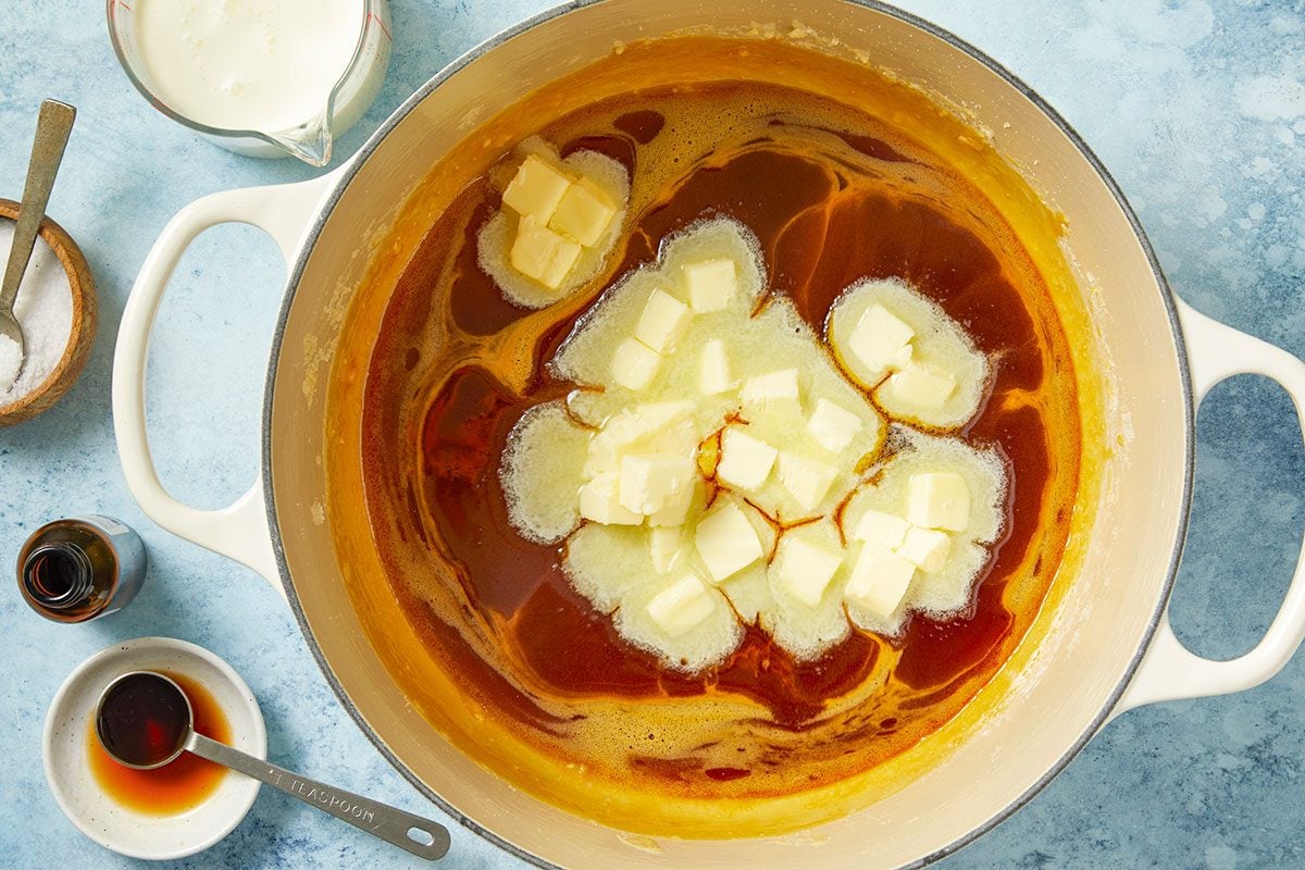 Overhead horizontal image of butter cubes being added to a saucepan with melted sugar. Also visible are cream, vanilla extract, and salt.