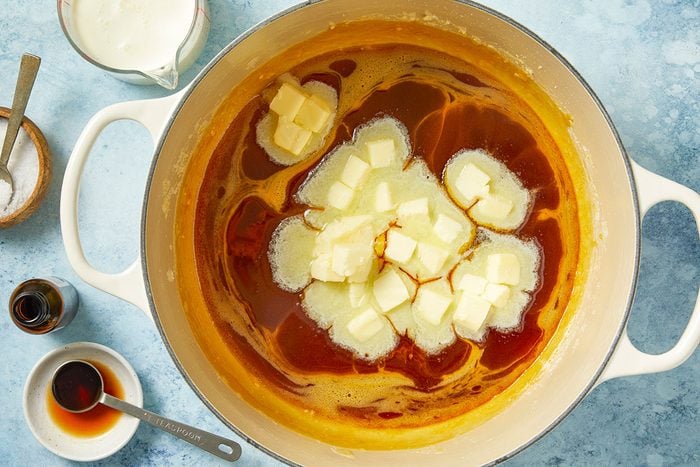 Overhead horizontal image of butter cubes being added to a saucepan with melted sugar. Also visible are cream, vanilla extract, and salt.