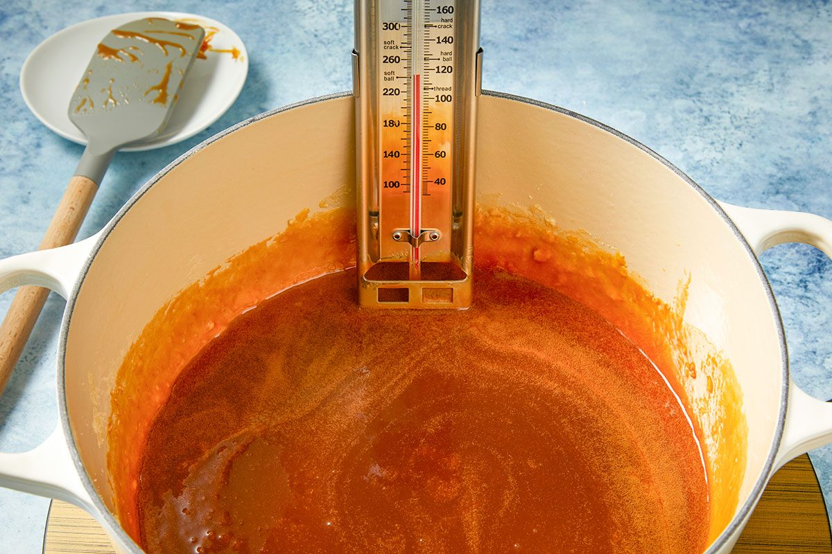 Eye Level horizontal image of caramel being cooked in a pot. A candy thermometer is clipped on the side of the pot. There is a spatula on a plate with some caramel on it.