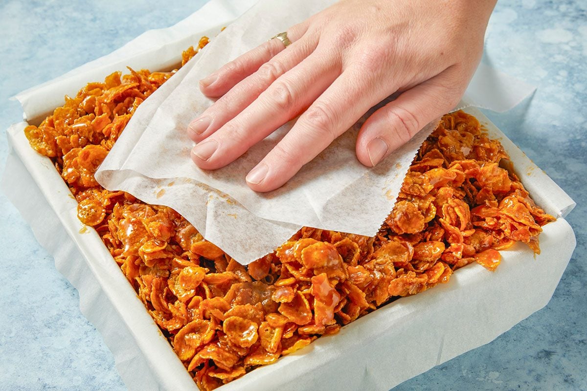 Eye Level horizontal image of cornflake cookies being pressed with parchment paper in a baking pan. The cookies are coated with a shiny syrup. A hand is pressing down on the parchment paper.