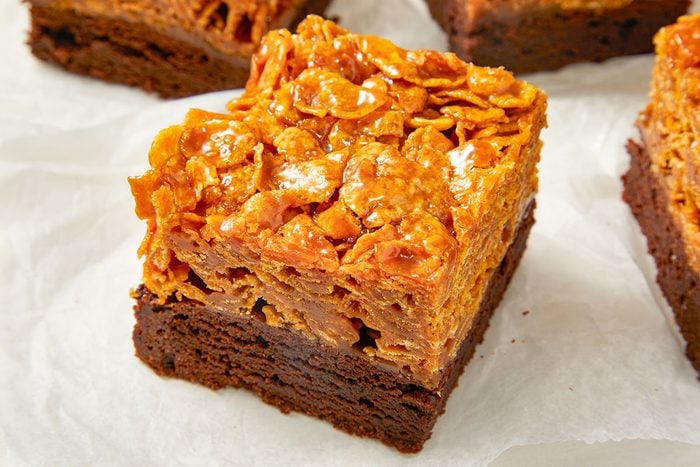 Eye Level horizontal image of a brownie square with a caramel cornflake topping on parchment paper. The brownie is a deep chocolate color with a slightly gooey texture and the topping has a bright caramel glaze with the individual cornflakes visible.