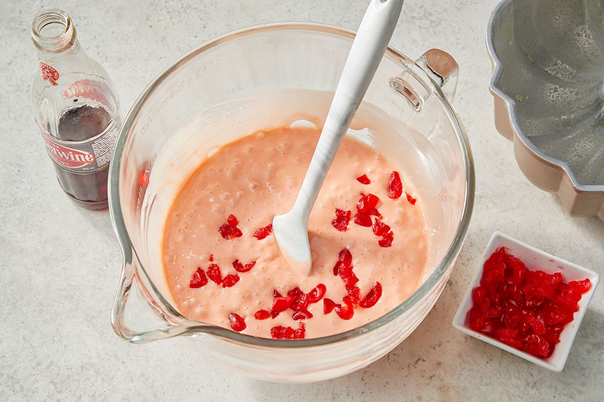 Overhead horizontal image of light pink batter in a clear glass mixing bowl. Chopped maraschino cherries are being stirred into the batter with a white spatula, with a bottle of Cheerwine soda and a greased bundt pan visible nearby.