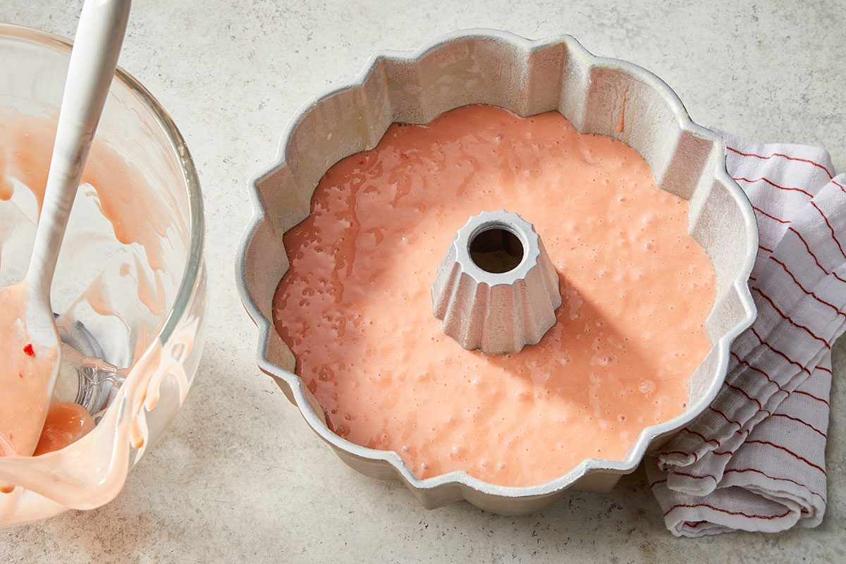 Overhead horizontal image of pink cake batter poured into a fluted gray bundt pan. A glass mixing bowl with residual batter and a spatula is on the left, while a white and red striped dishtowel rests on the right. This shot captures a step in the dessert's preparation.
