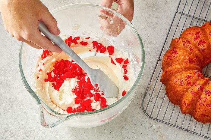 Eye Level horizontal image of hands mixing white frosting or filling with chopped red cherries in a clear glass bowl using a gray spatula. A freshly baked bundt cake with red cherries is cooling on a wire rack nearby.