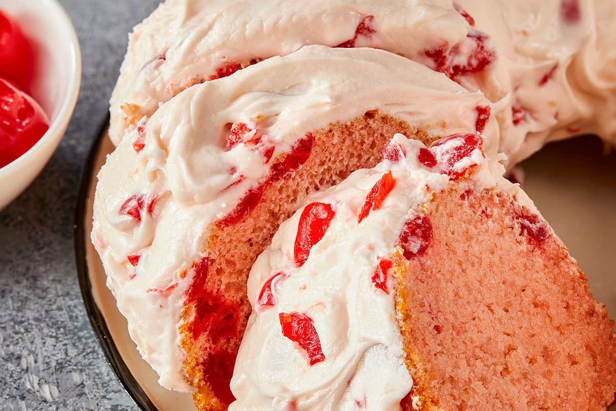 Eye Level horizontal image of several slices of a pink cake covered in thick white frosting. The cake and frosting are both speckled with bright red candied cherries or similar fruit pieces. A bowl with more red cherries is partially visible in the background.