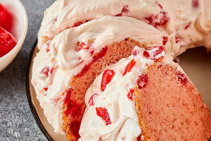 Eye Level horizontal image of several slices of a pink cake covered in thick white frosting. The cake and frosting are both speckled with bright red candied cherries or similar fruit pieces. A bowl with more red cherries is partially visible in the background.
