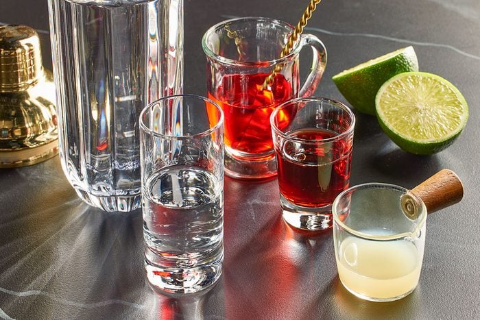 Eye Level horizontal image of cocktail ingredients set on a gray counter. There are 3 clear glasses with liquid in them. A cut lime is also in the frame.