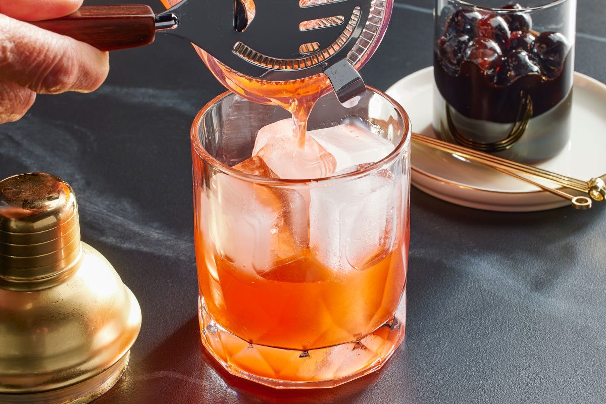 Eye Level horizontal image of an orange liquid being strained into a glass filled with ice. A shaker is visible to the left and a glass of cherries is visible in the background.